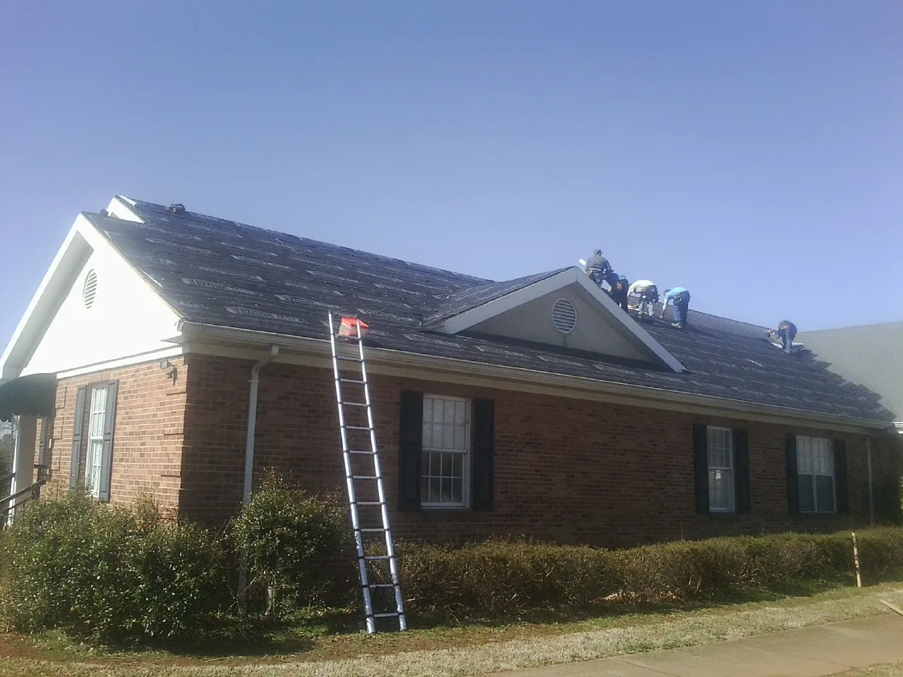 Crew tearing shingles off a brick ranch roof with a ladder set — Emergency Roof Repair work by our crew in Pierz
