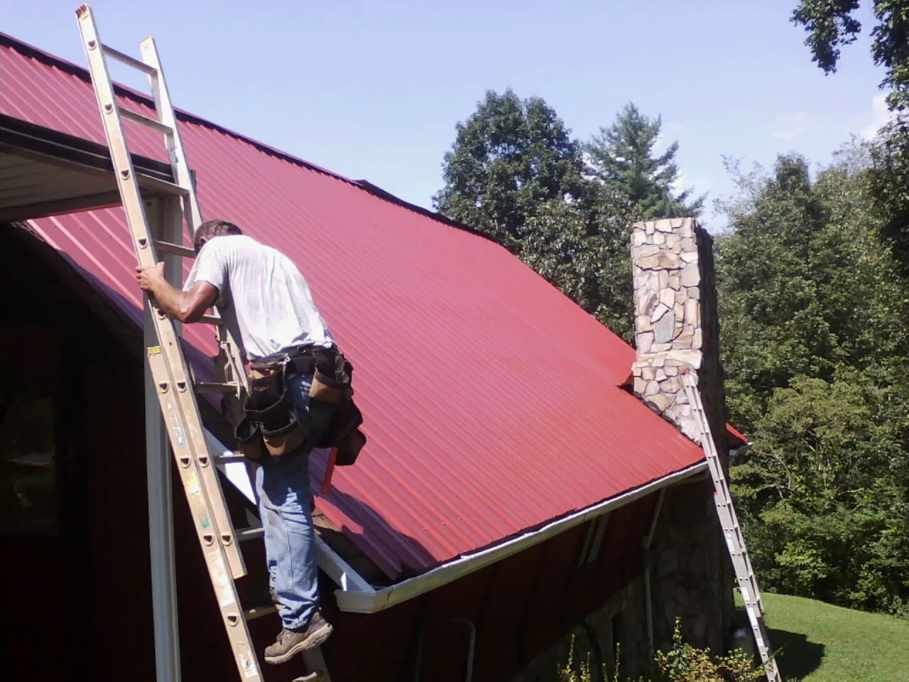 Roofer on a ladder beside a red metal roof and stone chimney — Soffit & Fascia Repair by Roofing Brothers in Pierz